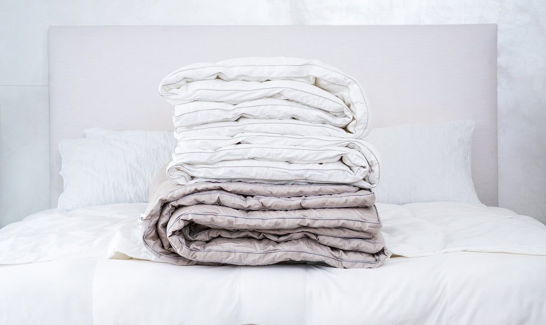 Two folded comforters stacked on a made bed, one white and one stone