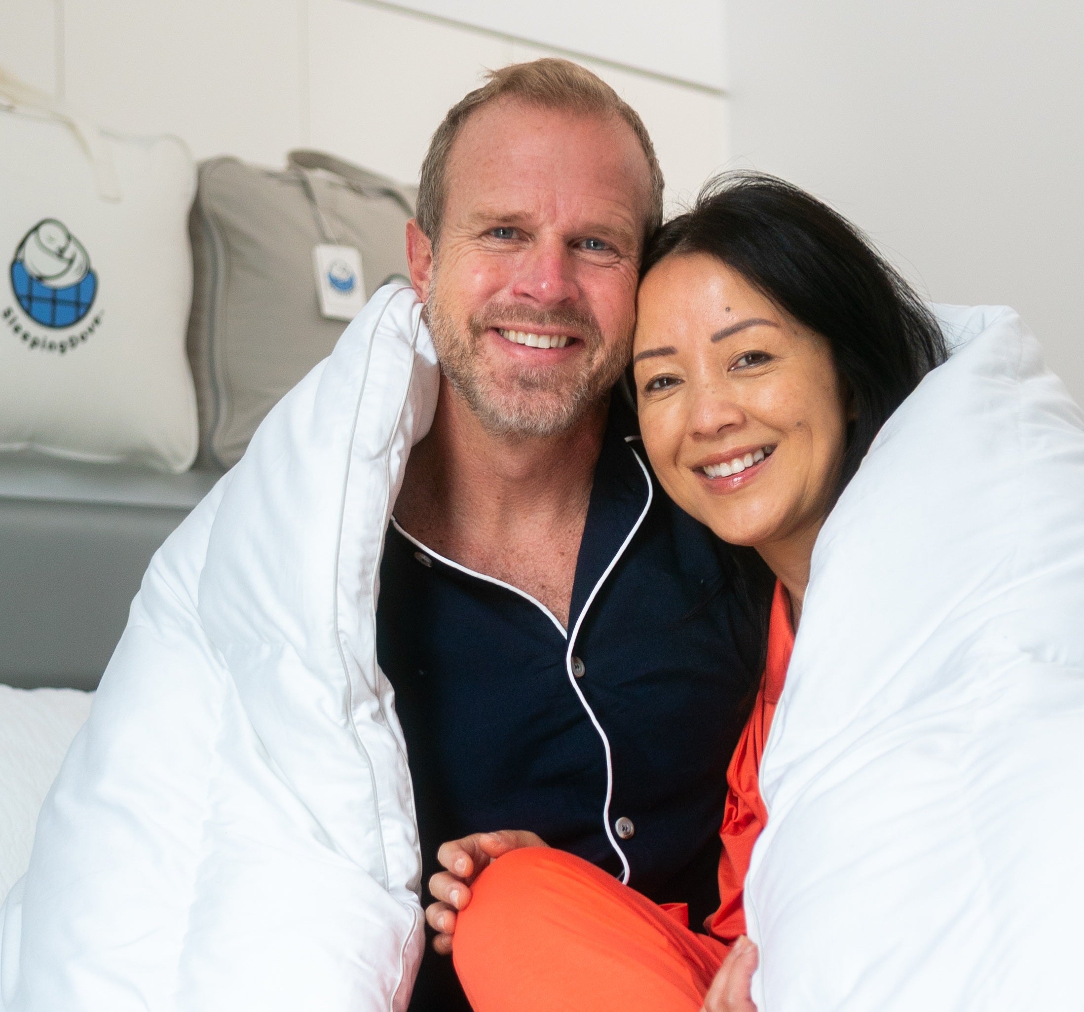 Man and woman hugging under a white Sleeping Dove comforter with a visible brand logo on a pillow.